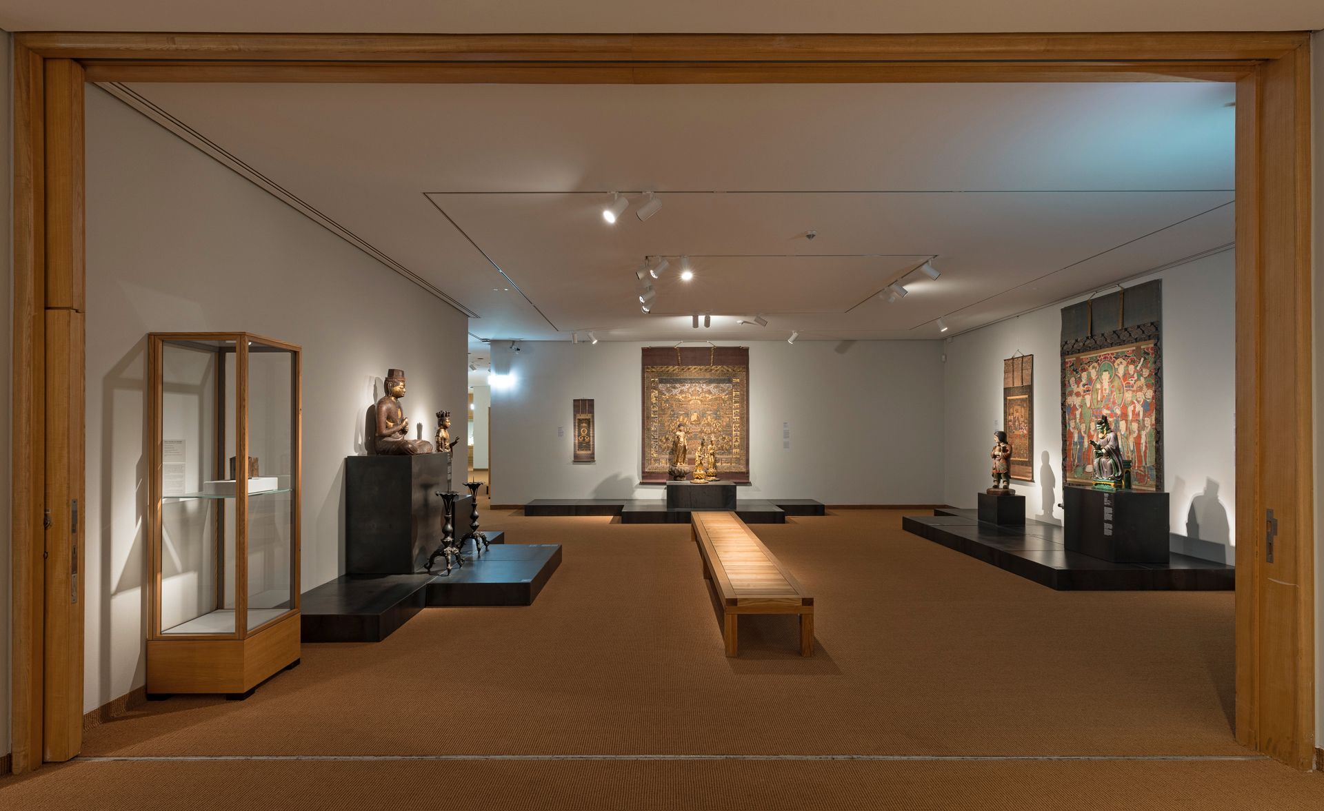 Blick in einen Ausstellungsraum im Museum für Ostasiatische Kunst mit buddhistischen Kunstwerken aus Ostasien, darunter Skulpturen, rituelle Objekte und Hängerollen