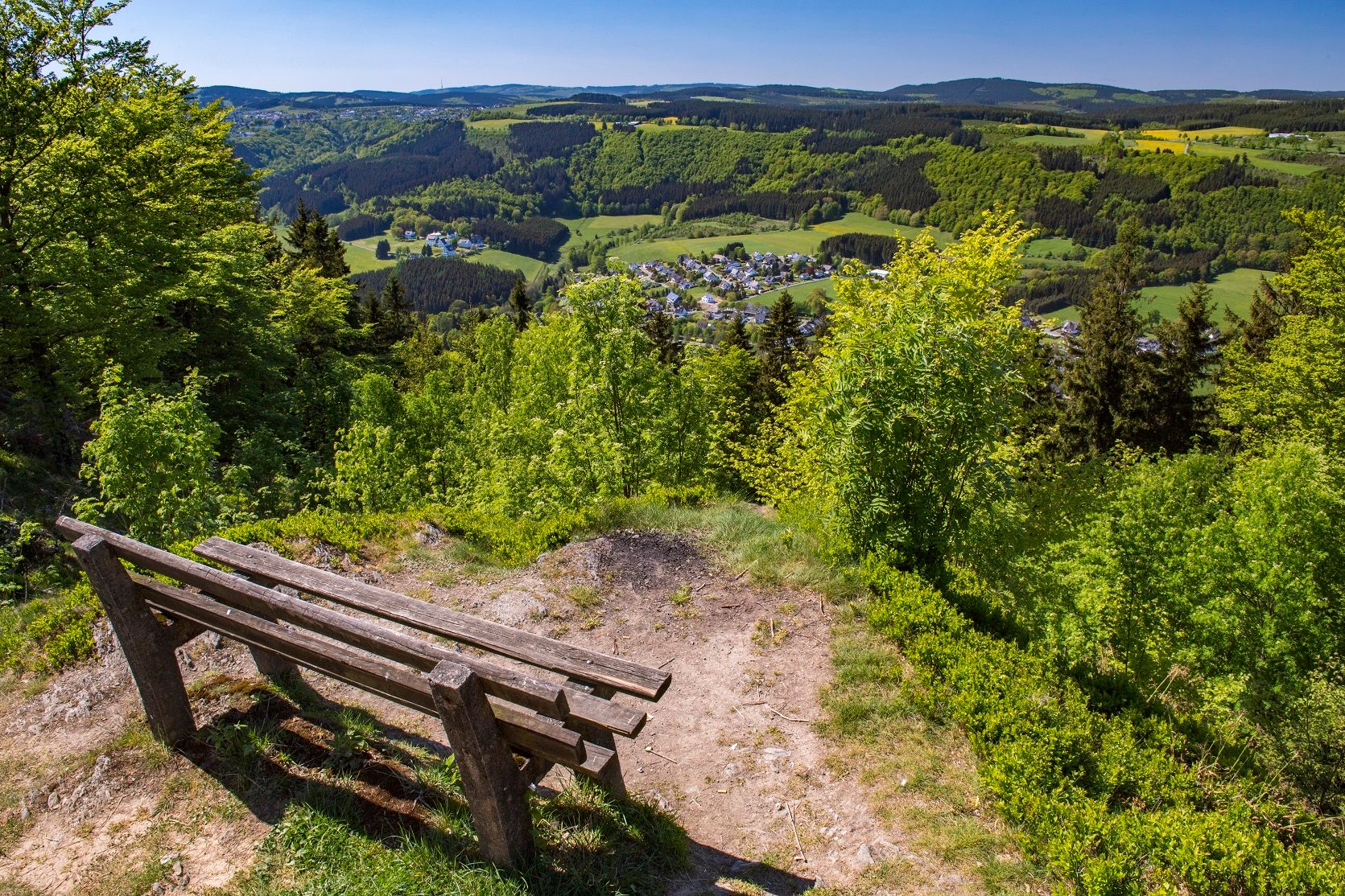 Holzbank auf einem Hügel mit Blick auf ein grünes Tal und ein Dorf.