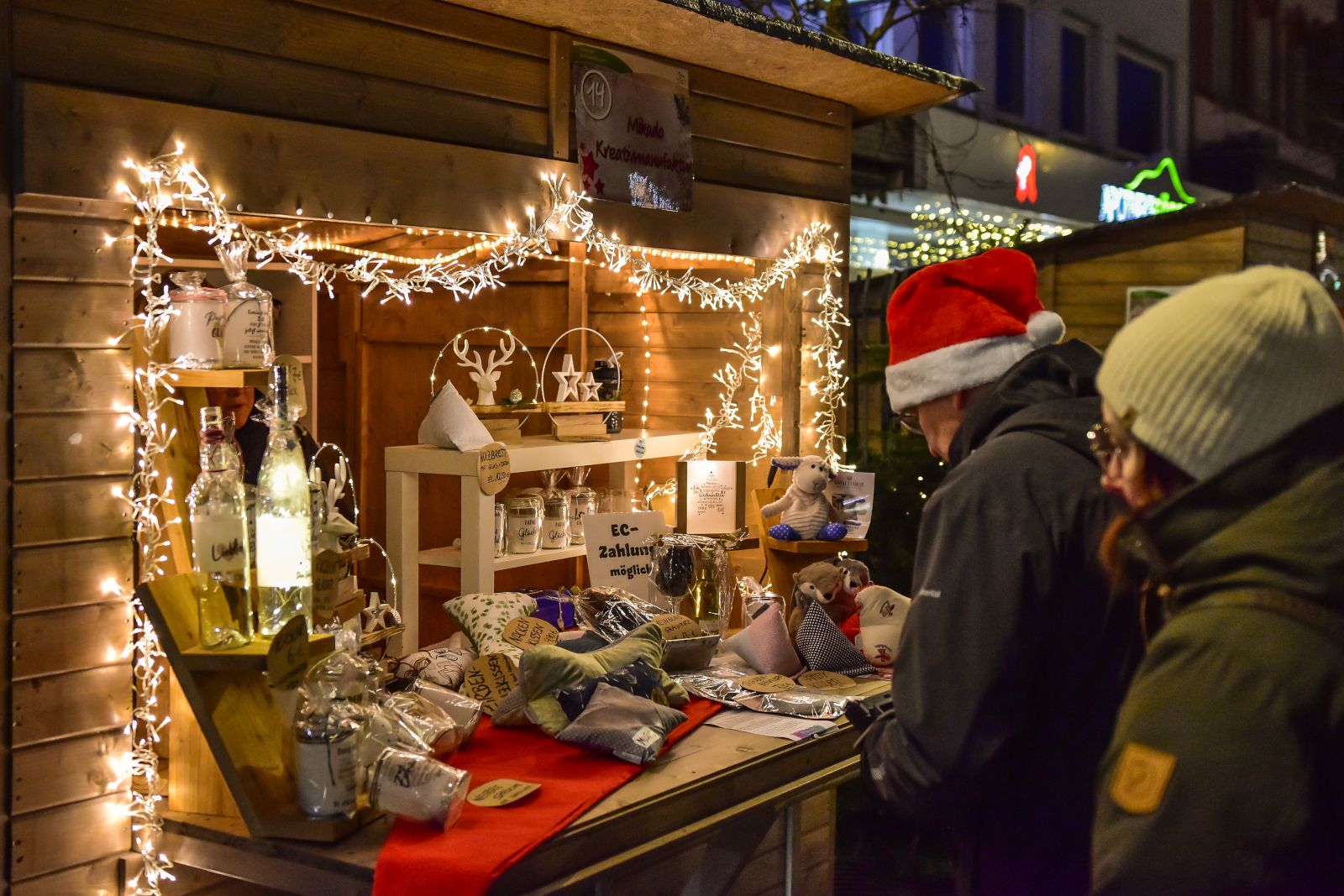 Weihnachtsmarkt Olsberg: beleuchteter Holzstand mit Deko und Geschenken, davor zwei Personen mit Mütze und Santa-Hut.