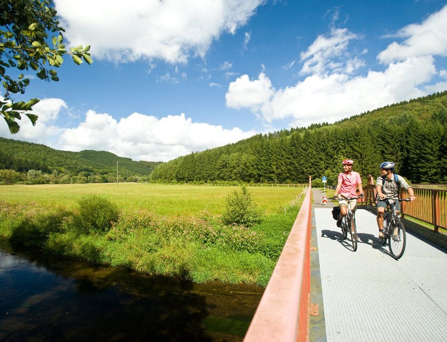 Zwei Radfahrer auf einer Brücke in der Eifel, umgeben von Wiesen und Wäldern.