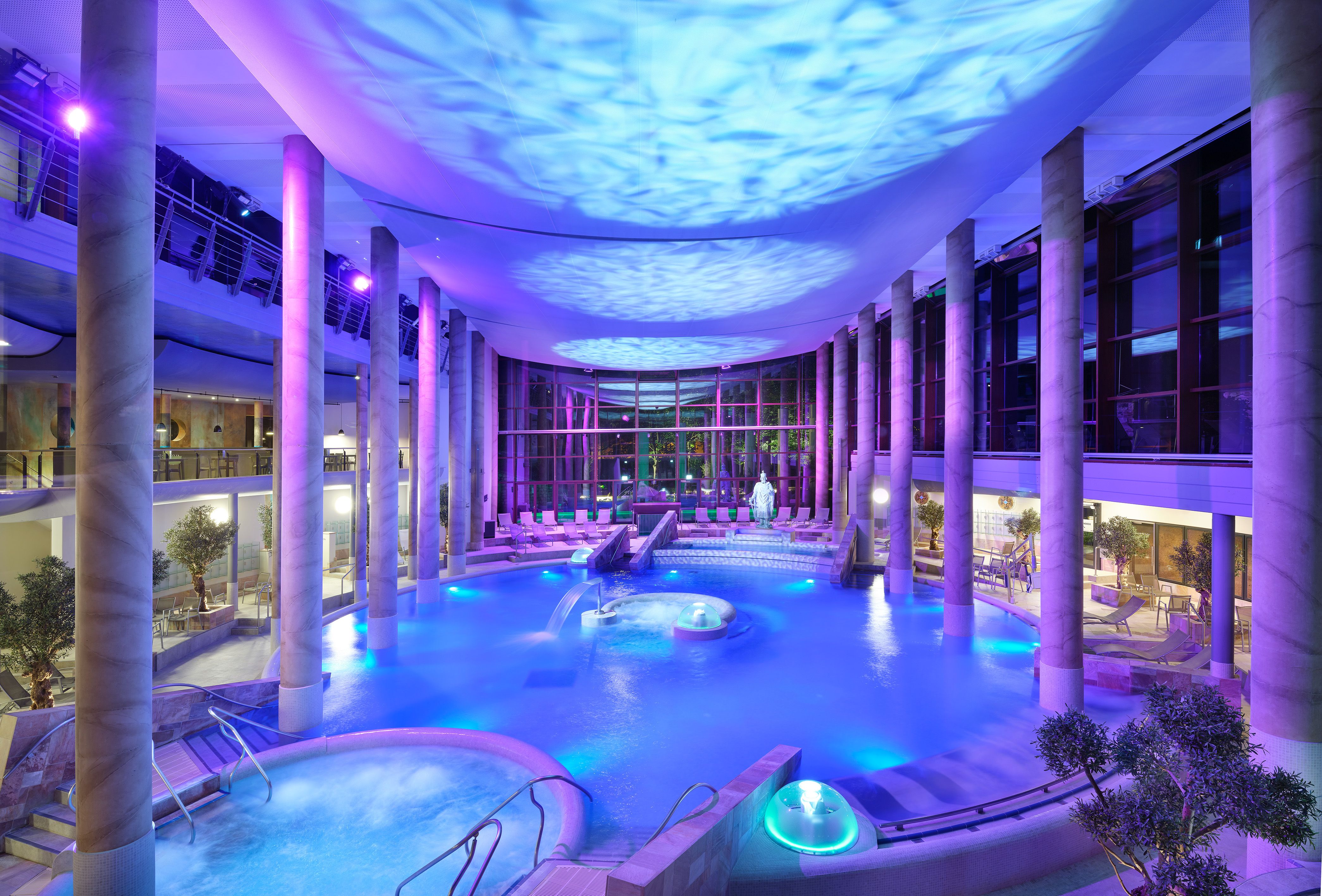 Innenansicht der Carolus Therme bei Nacht, beleuchtet in Blau- und Lilatönen. Der Innenbereich zeigt einen großen Pool, umgeben von Säulen und Liegestühlen.