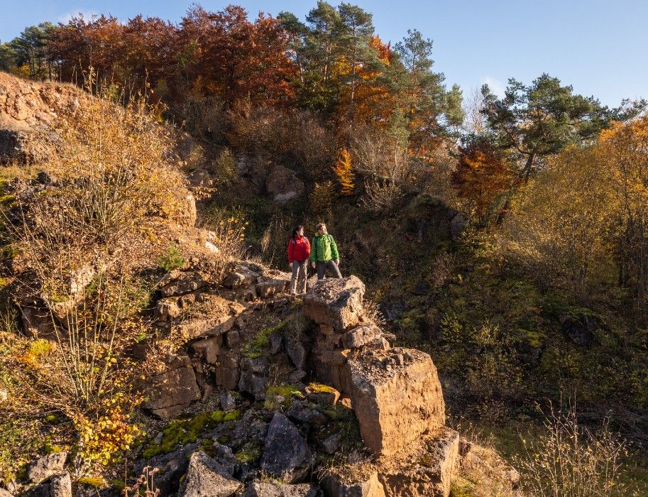 Zwei Wanderer stehen auf Felsen in einem herbstlichen Steinbruch in der Eifel.