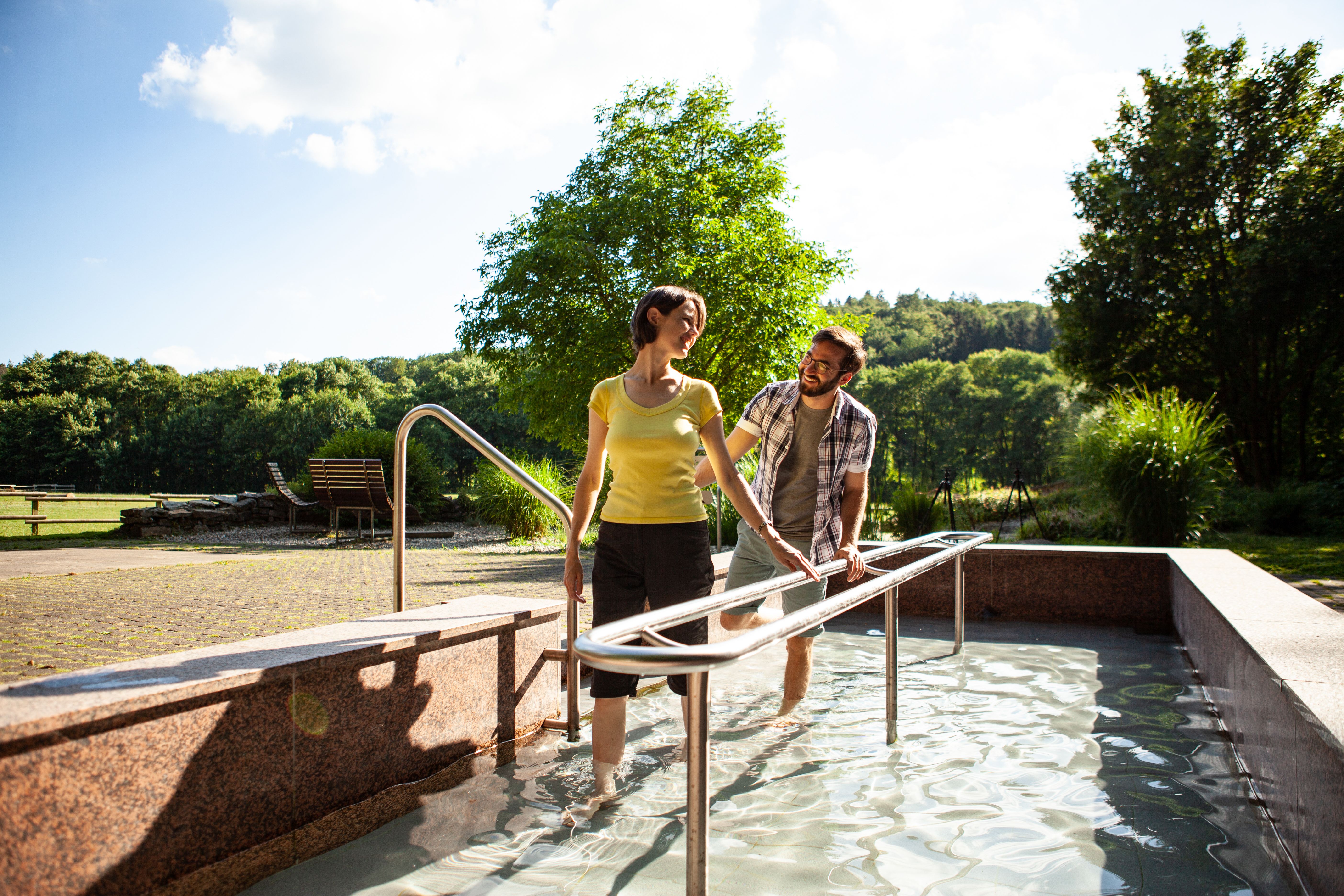 Zwei Personen gehen durch ein Kneippbecken im Freien, umgeben von Bäumen und Wiesen.