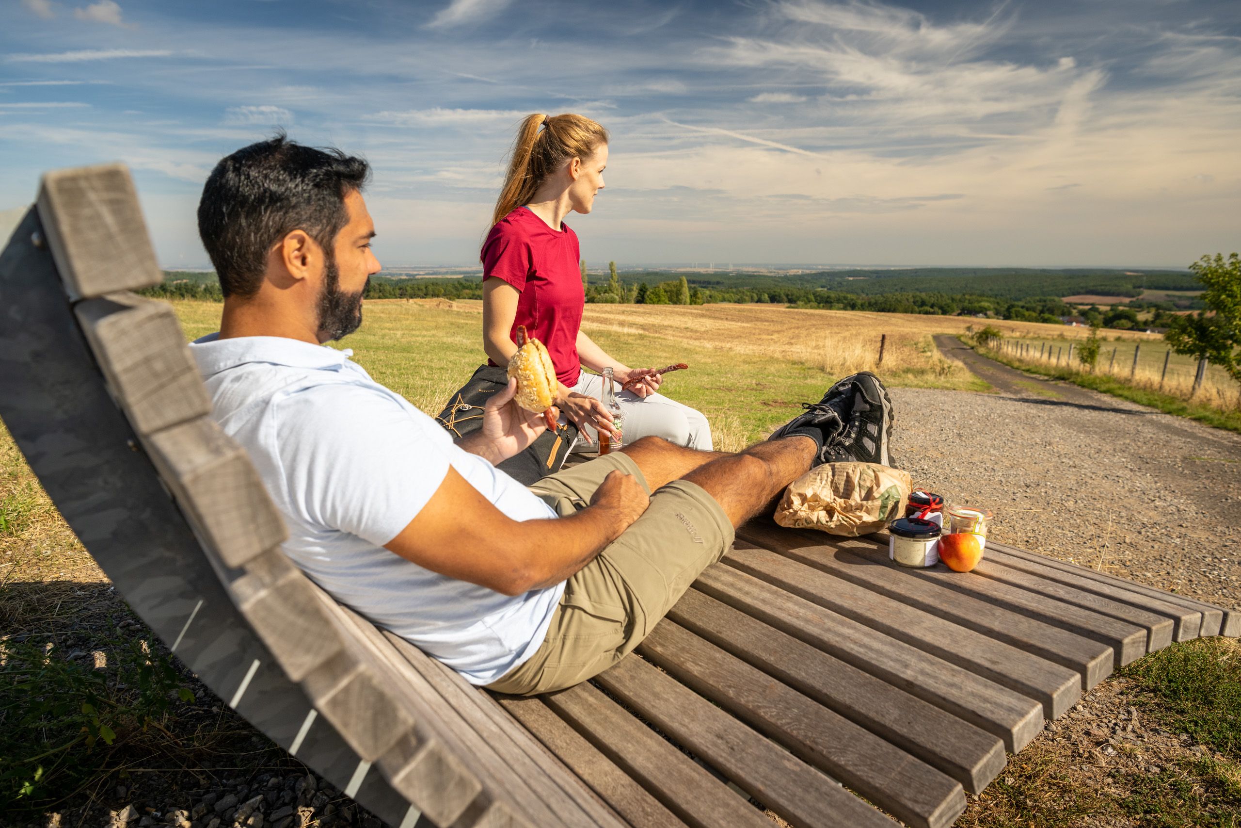 Zwei Personen entspannen auf einer Holzbank in einer weiten Landschaft. Sie genießen ein Picknick mit Sandwiches und Obst, während sie die Aussicht über Felder genießen.